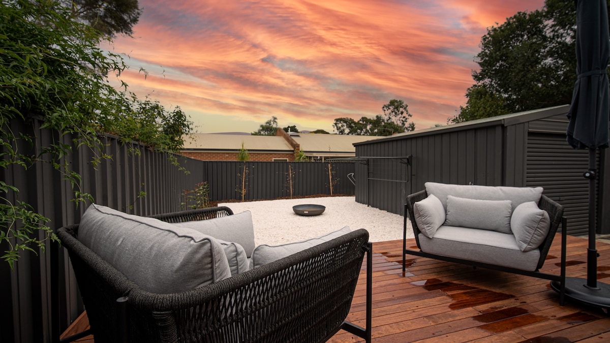 An outdoor deck area features two comfortable seating options with plush cushions. A firepit is centrally positioned on a gravel surface, surrounded by tall privacy screens. Lush greenery is visible along the edges, complementing the vibrant sunset sky above.