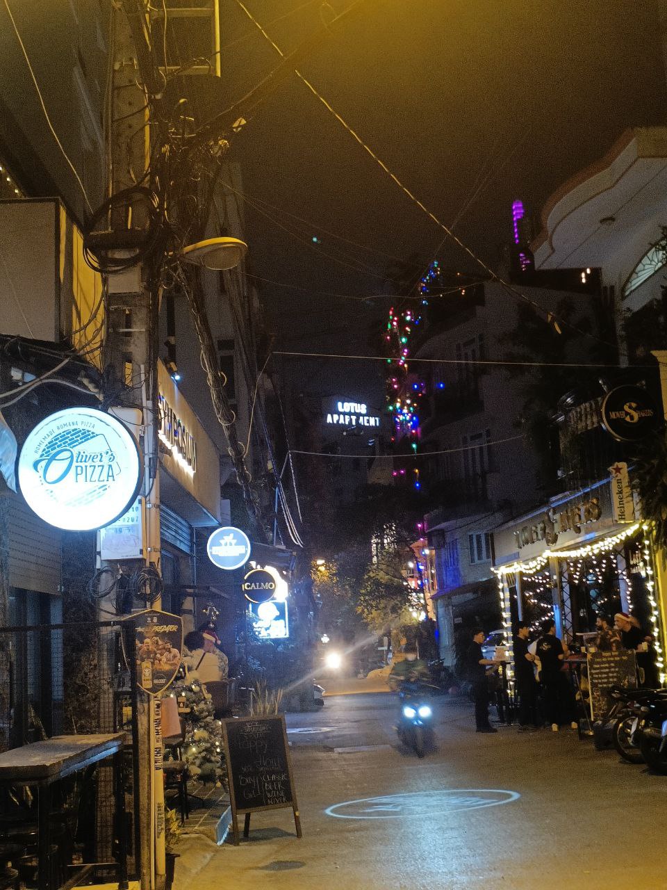 A narrow street illuminated by warm lights sets a lively atmosphere. Unique signage from local eateries is visible, including a pizzeria. Motorcycles are parked, and people can be seen enjoying the evening at outdoor dining areas, with a building featuring a name illuminated in the background.