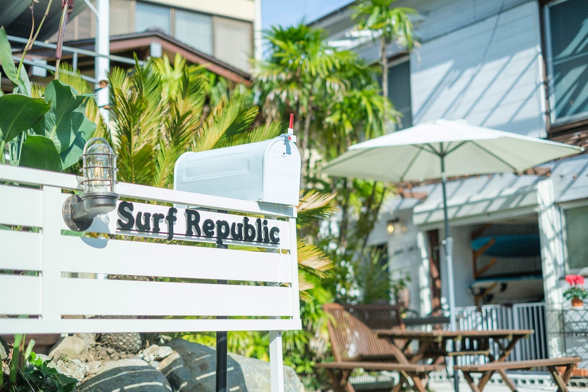 The outdoor area displays a welcoming sign labeled 'Surf Republic' beside a mailbox. A patio table with an umbrella and wooden seating creates a relaxed atmosphere. Lush greenery surrounds the setting, enhancing the overall appeal.