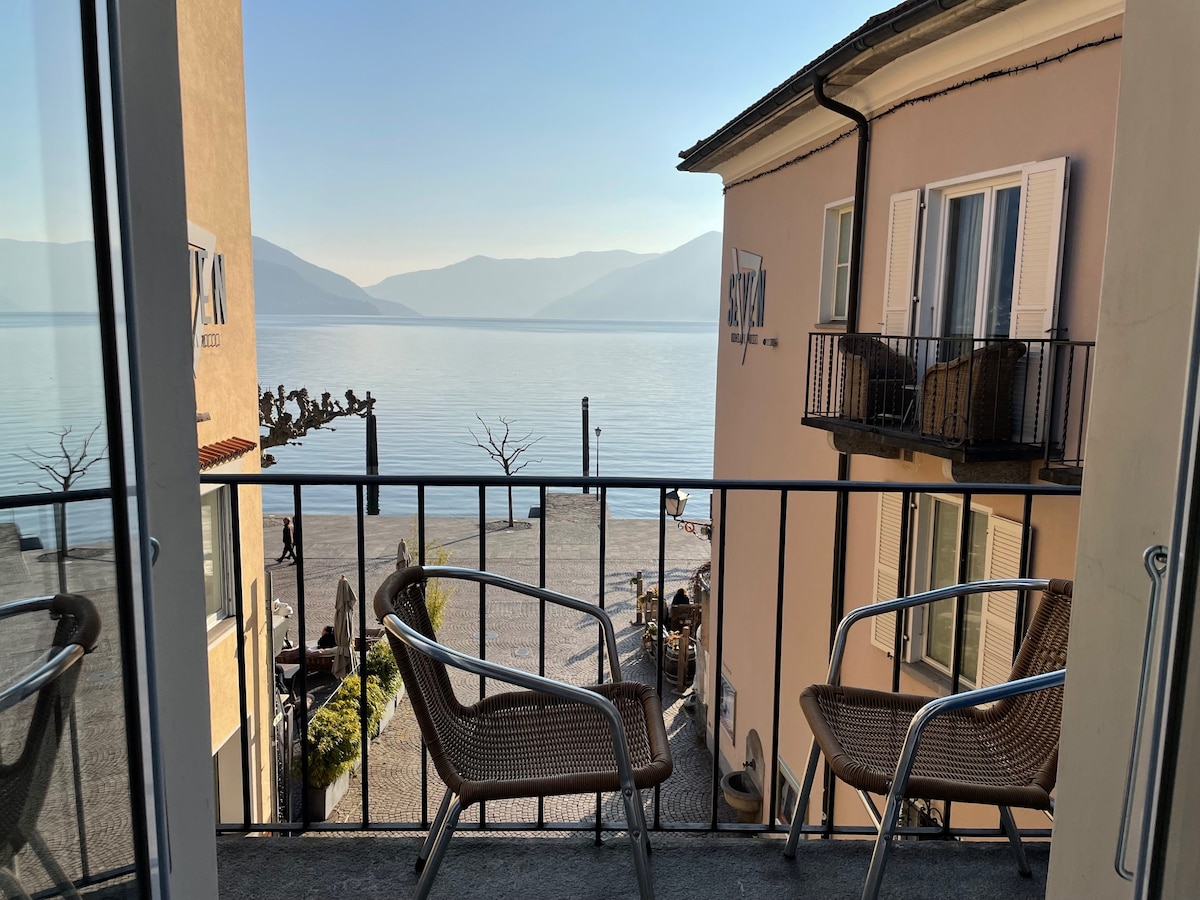 A balcony view reveals a calm lake surrounded by mountains, with two wicker chairs positioned along the railing. Soft morning light enhances the tranquil scene, inviting relaxation by the water's edge.