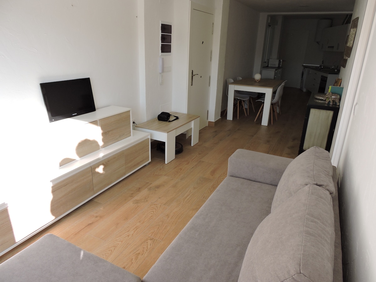 A light-filled living area is visible, showcasing a gray sofa and minimalistic furniture. A flat-screen TV is placed on a low cabinet. A dining table with chairs is observed in the background, along with a glimpse of the kitchen area, enhancing the open layout.