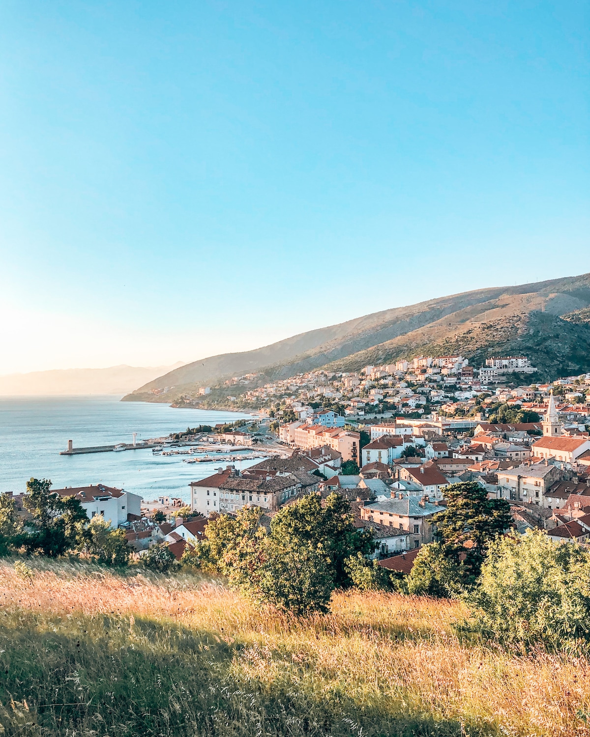 An expansive view of Senj is captured, revealing a coastal town nestled at the foot of rolling hills. The harbor is visible with boats docked, while the charming homes with terracotta roofs stretch toward the sea, under a clear blue sky.