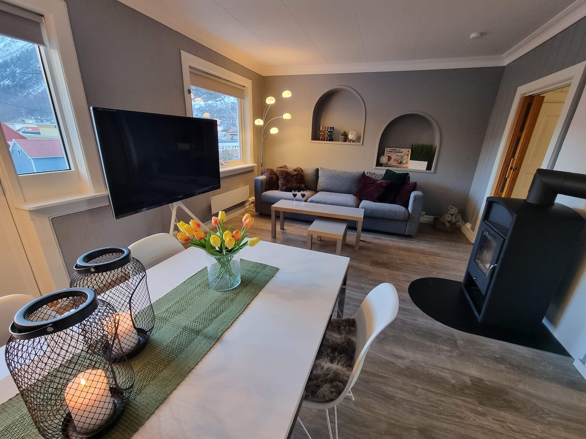 A bright living area features a dining table set with a green runner and a vase of tulips. A comfortable sofa and decorative niches add charm. A 60" television stands on a media unit, while an electric stove enhances the space's warmth.