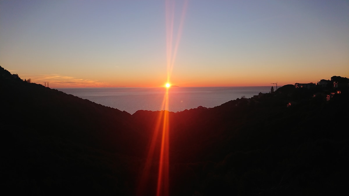 A panoramic view captures the sunset over the sea, with golden rays reflecting on the water. Silhouettes of hills create a natural frame around the horizon, while distant lights from nearby houses twinkle softly in the fading light.