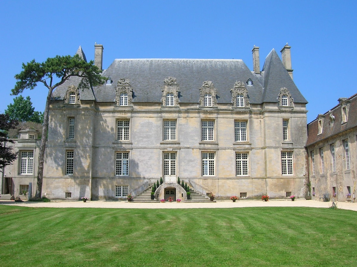 The stately exterior of the castle is highlighted by intricate stonework and large, symmetrically arranged windows. A well-maintained lawn leads to the main entrance, framed by steps and flowerbeds. The clear blue sky provides a bright backdrop to this historic structure.