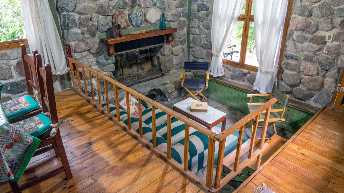 A sunlit living area features a stone fireplace as the focal point, surrounded by rustic wooden accents. A wooden barrier defines the sunken seating area, which includes a striped sofa and a small table. Natural light filters in through large windows dressed with white curtains.