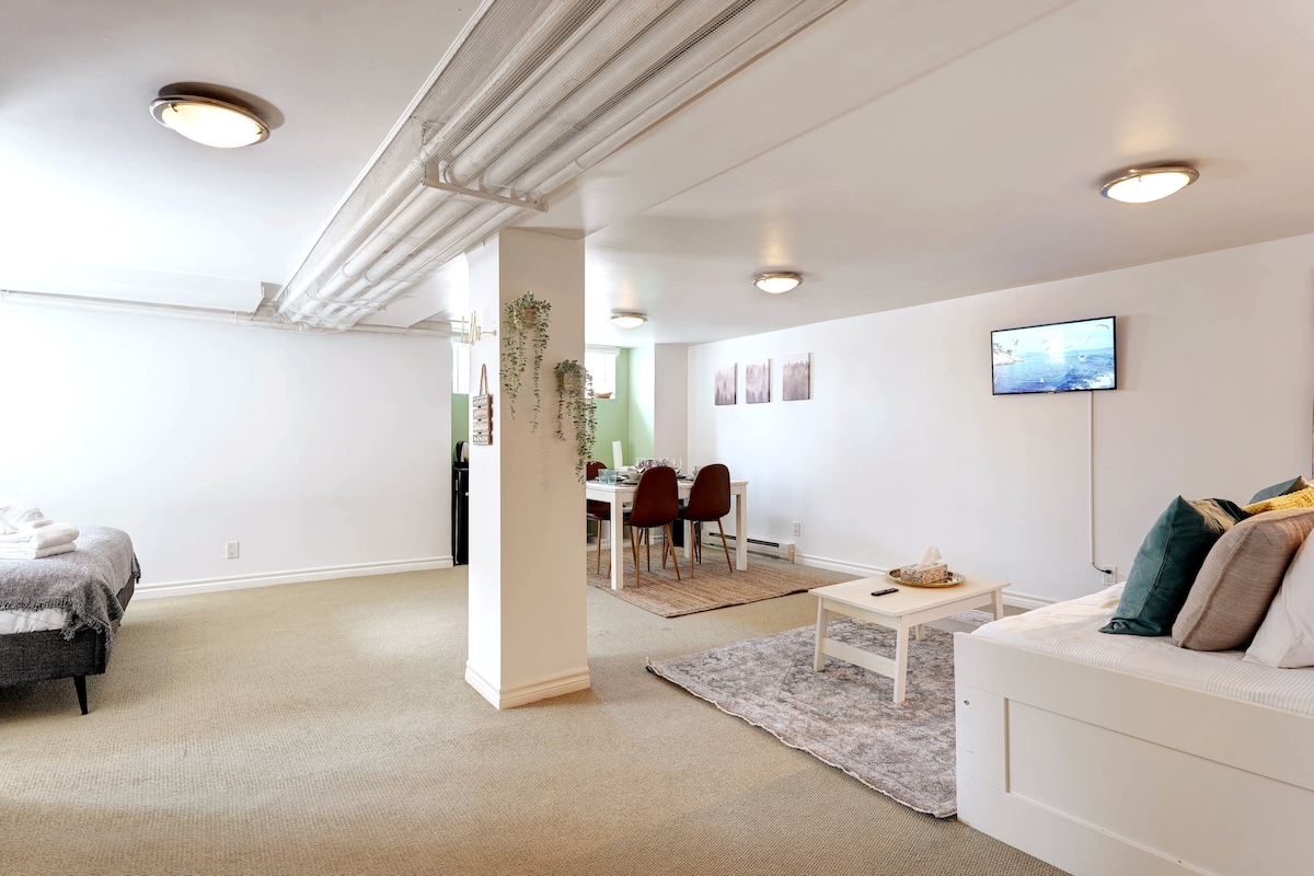 The spacious studio features a well-lit layout with a comfortable seating area and a convenient workspace. A queen bed is positioned alongside a king-size sofa bed. A dining area with four chairs is visible, complemented by a television mounted on the wall.