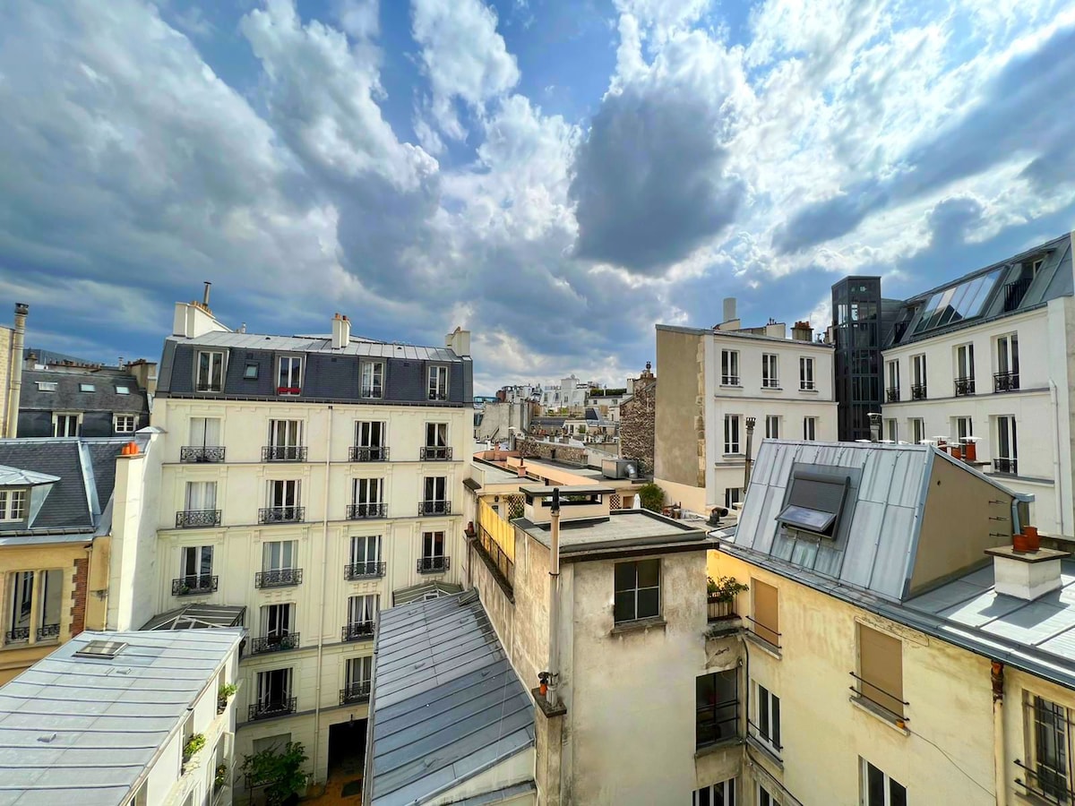 The image presents a panoramic view of Parisian rooftops, characterized by a mix of architectural styles. Various buildings with flat and sloped roofs are visible, alongside hints of greenery peeking through. The sky is filled with dynamic clouds, suggesting a change in weather.