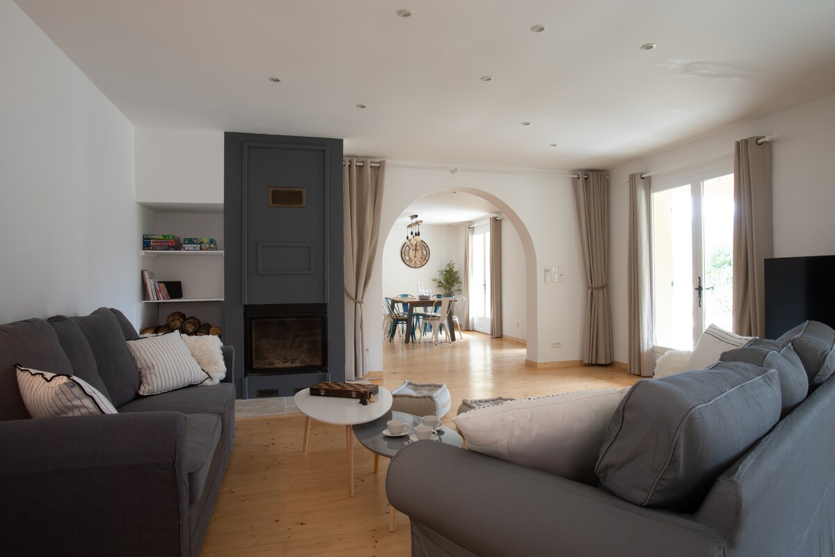 A spacious living area features two comfortable sofas oriented towards a coffee table. A modern fireplace is set into a stylish grey wall. Natural light filters through large windows, with light curtains framing the view. An archway leads to a dining space visible in the background.