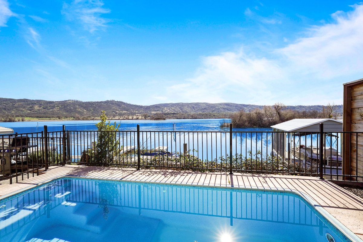 A crystal-clear sports pool is framed by a black fence, offering views of a tranquil lake and surrounding hills. Bright blue skies reflect on the water's surface, while the covered dock and lush greenery add to the serene setting.