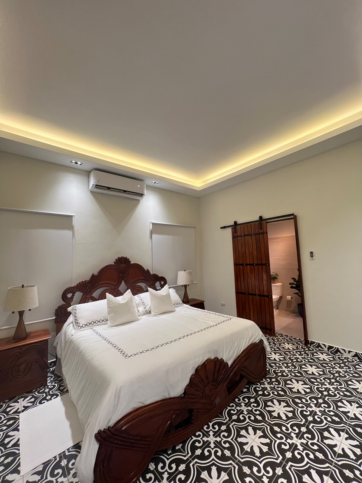 A spacious bedroom features a wooden headboard and is lit by soft overhead lighting. The floor is adorned with decorative tiles, while two bedside tables hold lamps. A sliding barn door leads to a bathroom area, enhancing the room's overall functionality.