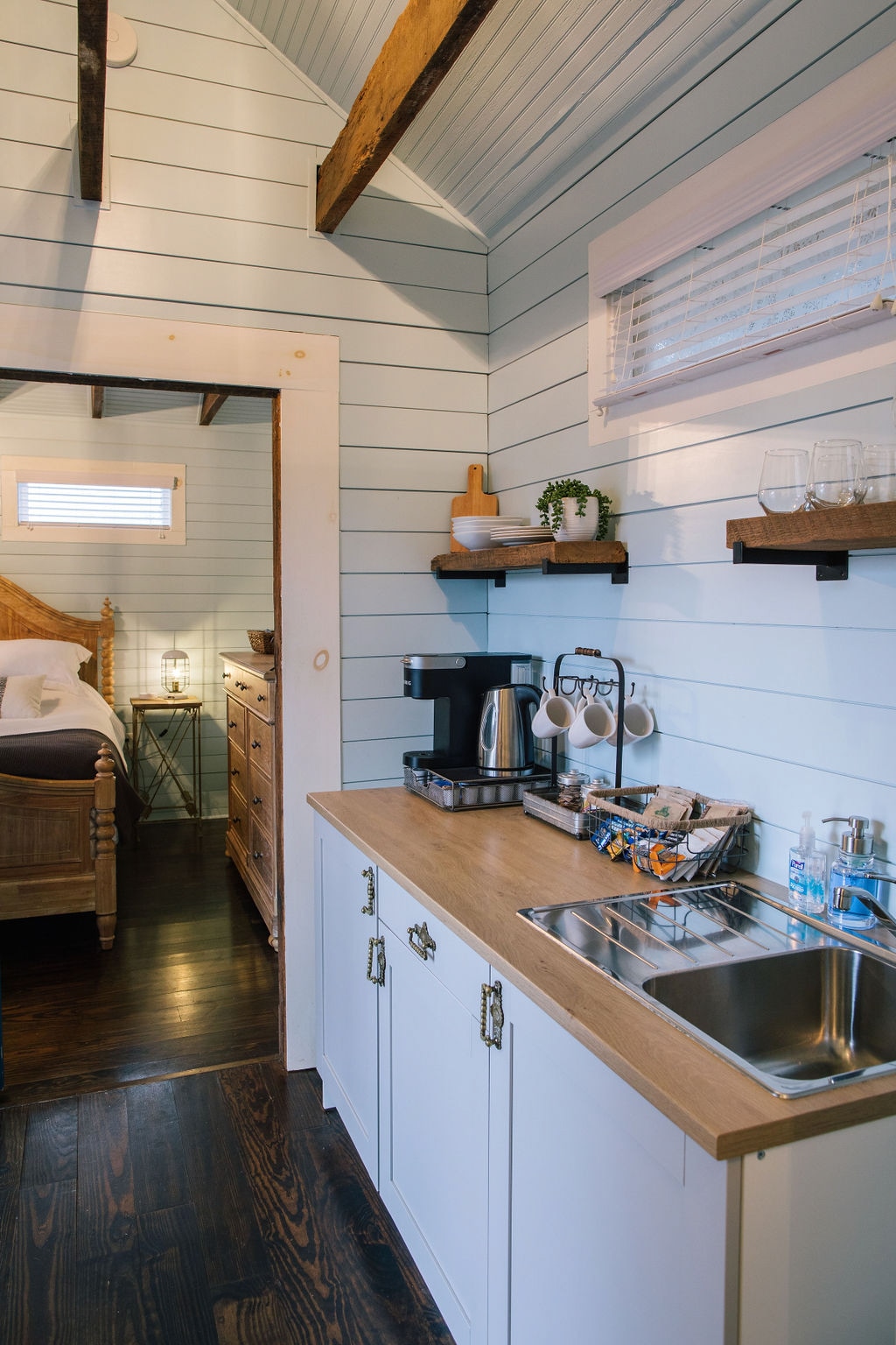 The kitchenette features a wood countertop with a coffee maker, tea kettle, and mini fridge. Open shelves display glassware and kitchen essentials. Light blue walls enhance the space, while a glimpse of the cozy sleeping area is visible through the doorway.