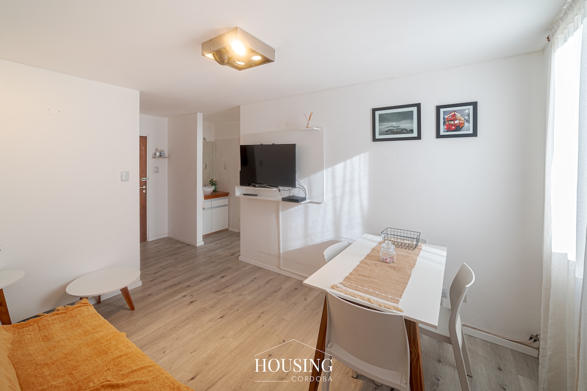 An inviting living area features a light wooden floor and a white wall with two framed artworks. A television is mounted on a white unit, while a dining table with four chairs is positioned nearby. Natural light fills the space, complemented by stylish ceiling lighting.