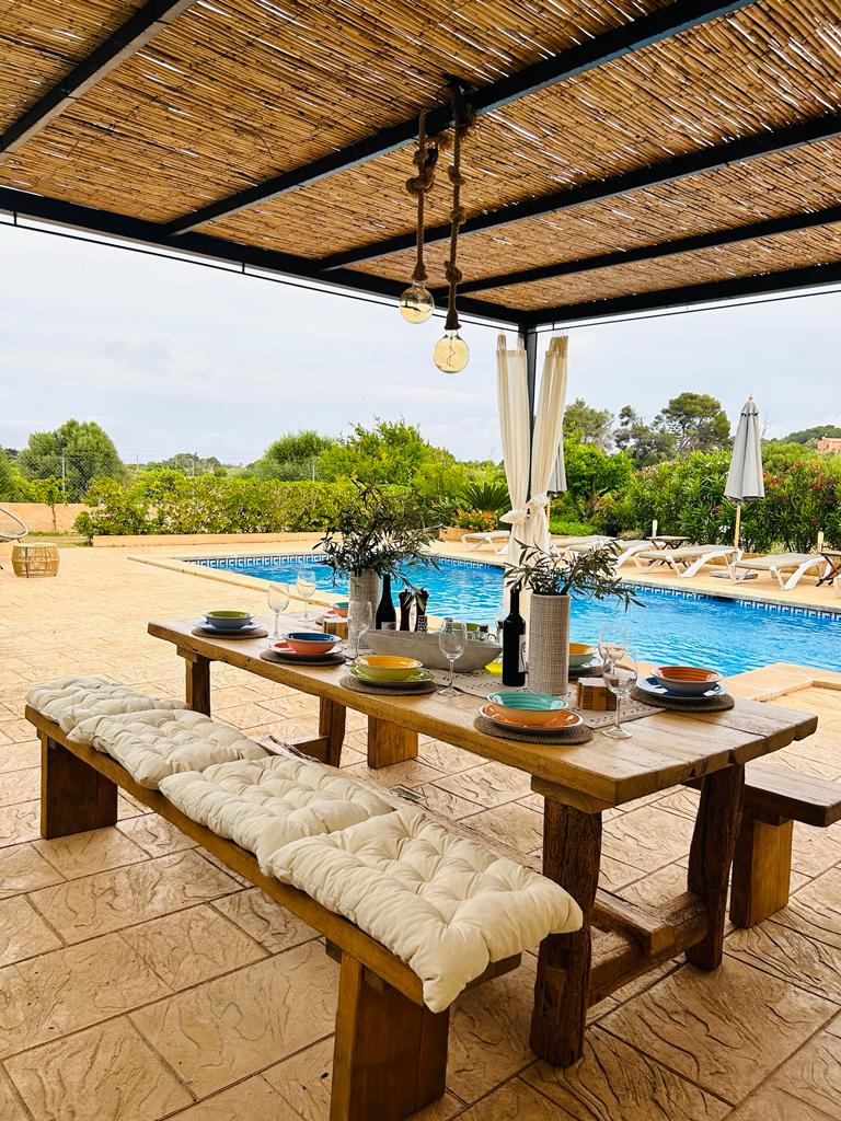 An outdoor dining area features a rustic wooden table adorned with colorful dishware and glassware. A shaded pergola provides shelter above, while a refreshing pool glistens nearby. Sun loungers are arranged beside the pool, set against a backdrop of lush greenery.