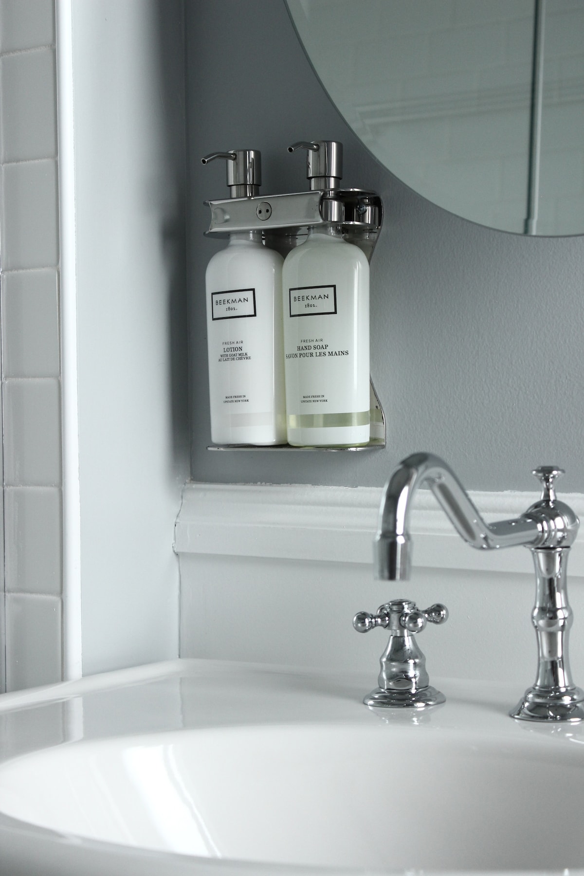 A clean and well-maintained bathroom sink is shown, featuring a polished faucet. Beside the sink, a wall-mounted dispenser holds complimentary toiletries, including two bottles of liquid soap and lotion. A round mirror is positioned above the sink, reflecting the simple and elegant design.