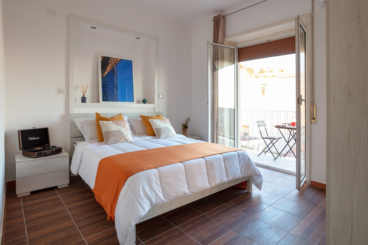 A bright bedroom is presented with a spacious bed featuring an orange throw blanket. Large glass doors open to a small balcony with two chairs and a table. Natural light fills the room, complemented by light-colored walls and warm wooden flooring.