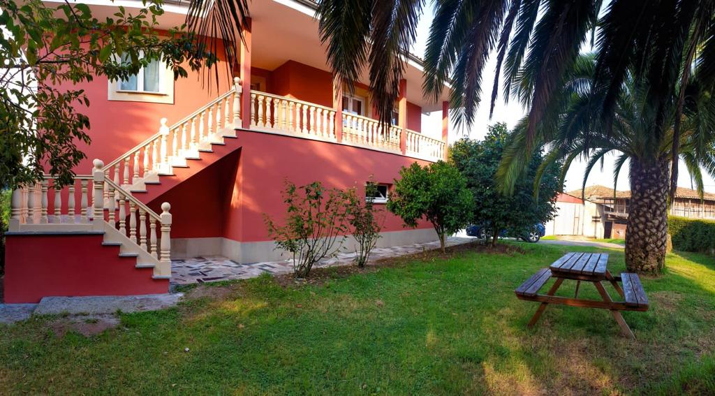 The exterior of a pink two-story house is shown, featuring a decorative staircase leading to the upper level. A grassy area with a wooden picnic table is visible, surrounded by palm trees and shrubbery, creating a serene outdoor environment.