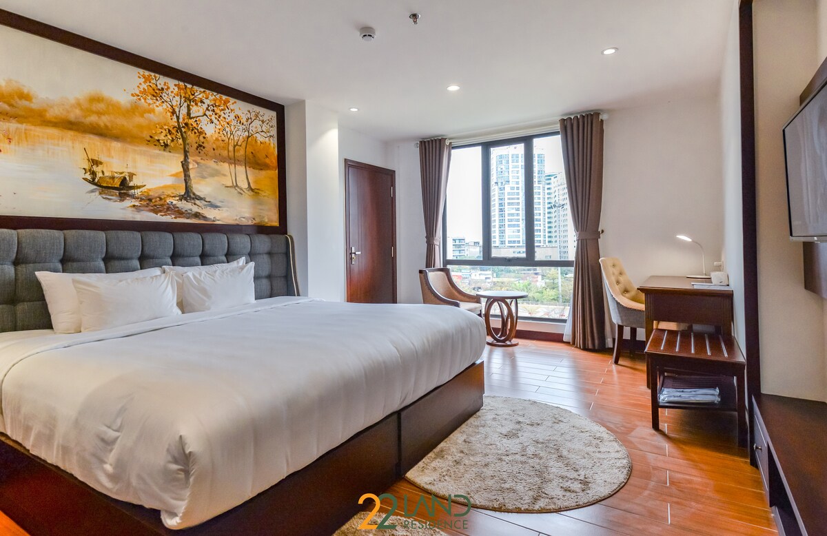 A spacious bedroom features a large bed with a gray tufted headboard. Artwork depicting a serene landscape adorns the wall. Natural light fills the room through a large window with sheer curtains, and a small workspace with a chair is visible near the window.