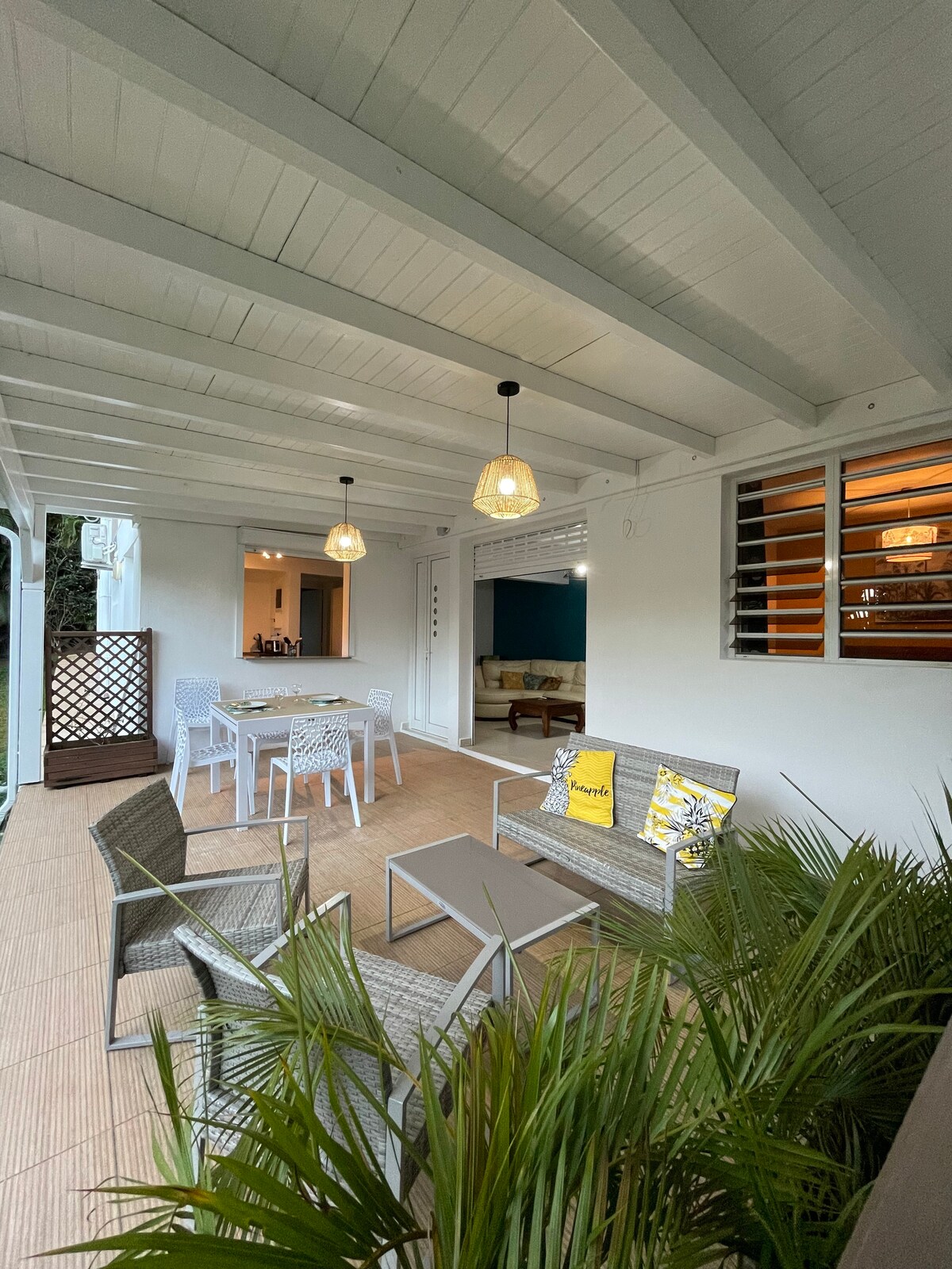 An outdoor terrace features a combination of lounge and dining spaces, furnished with modern chairs and a small table. Pendant lighting hangs down from a white ceiling, and lush greenery surrounds the area, creating a tranquil and inviting setting.
