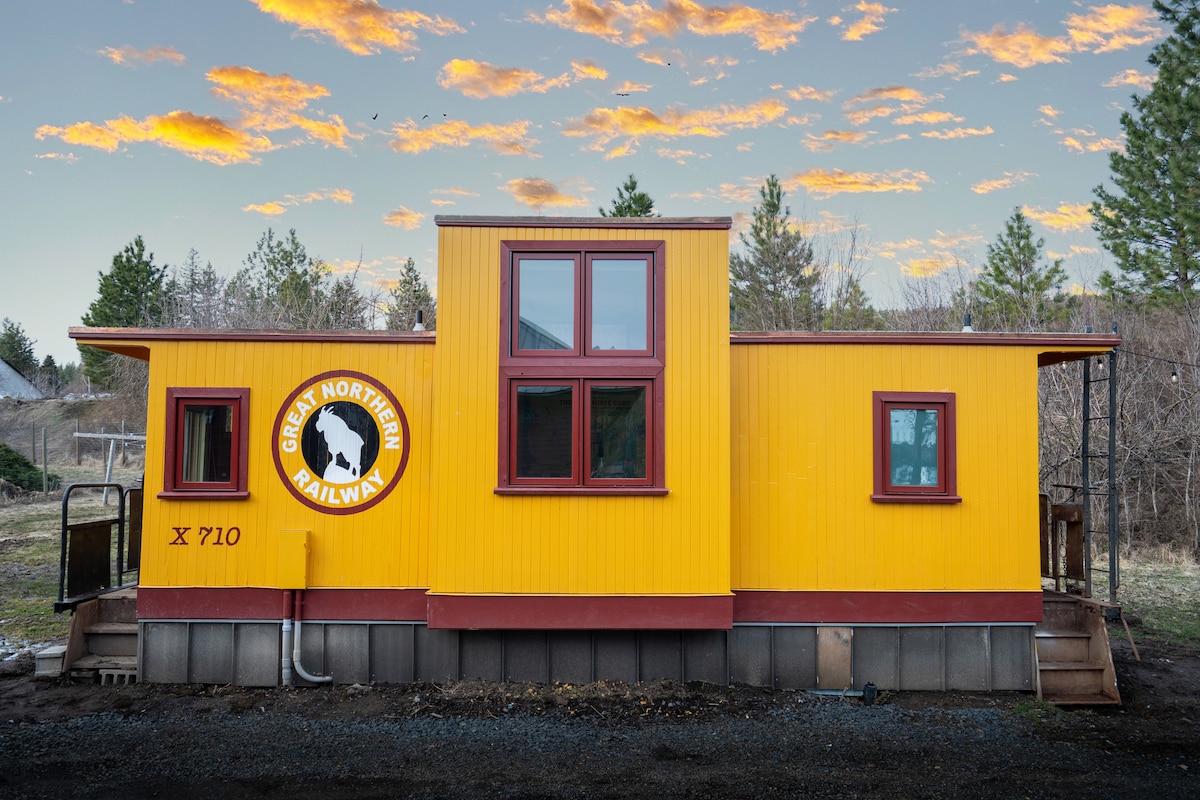 A charming train car exterior is presented, featuring bright yellow siding with red accents. Large windows allow natural light to enter. The Great Northern Railway logo is displayed prominently. Surrounding greenery and a colorful sky enhance the scenic backdrop.
