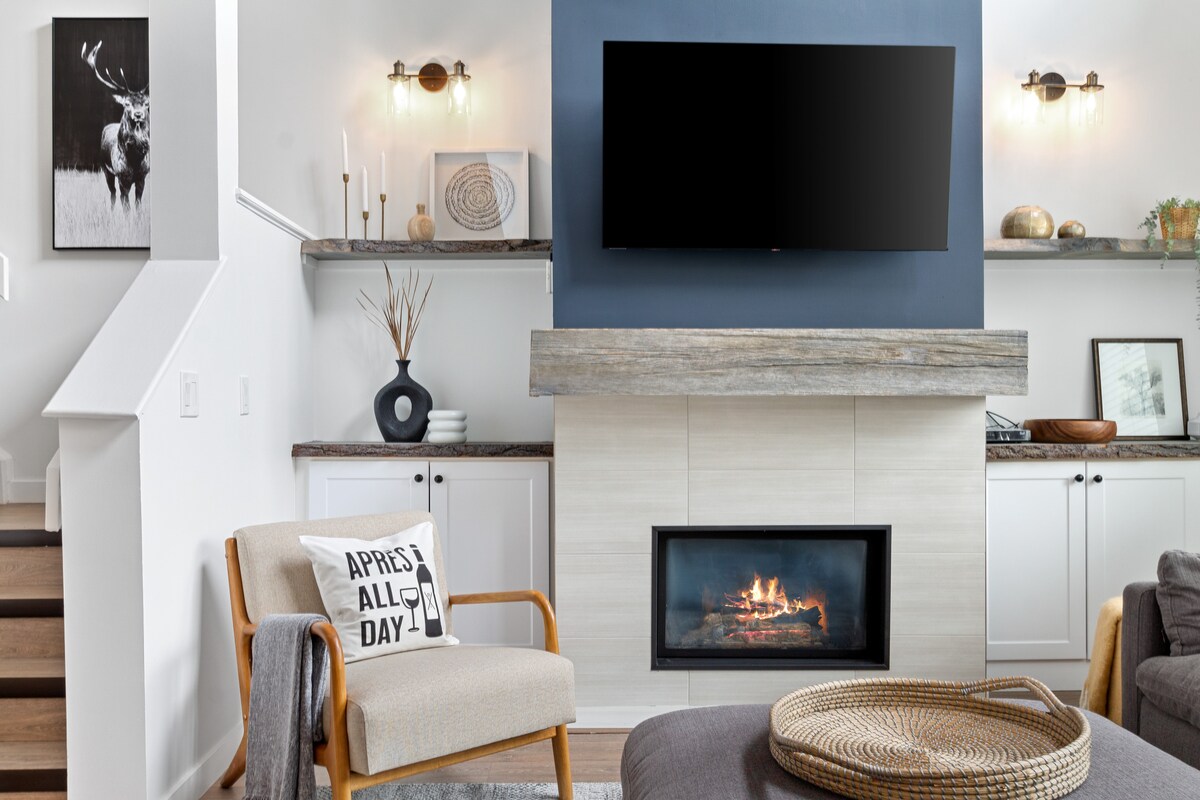 A modern living room features a stone fireplace with a sleek wooden mantle, topped by a flat-screen TV. Shelves on either side display decorative items and art. A comfortable chair with a cushion and a woven basket complete the cozy setting, illuminated by soft lighting.