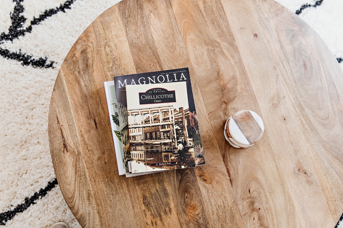 A wooden coffee table features a magazine about Chillicothe, Ohio, prominently displayed. A decorative stone object rests beside the magazine. The table's surface showcases a natural wood grain pattern, complementing the soft textures of a patterned rug underneath.