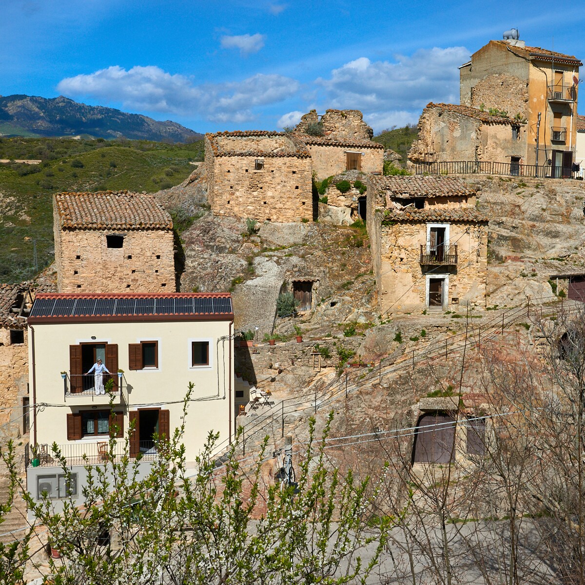 A scenic view showcases a blend of ancient stone buildings and a modern cottage. The cottage features solar panels on the roof and a balcony overlooking the surrounding landscape, while remnants of historic structures sit atop rocky terrain beneath a clear blue sky.