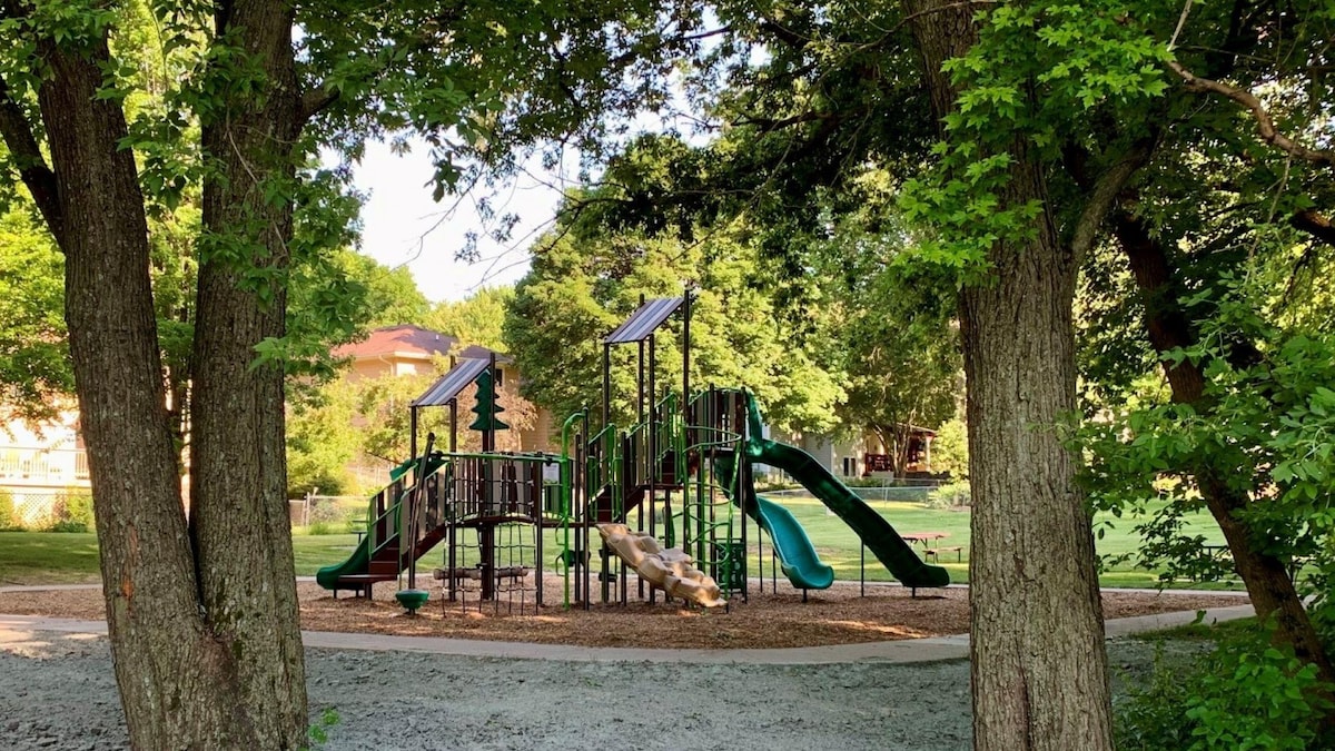 A community playground is surrounded by tall trees, featuring multiple slides and climbing structures. Soft mulch covers the ground below the play area, with open green space visible in the background. Bright sunlight filters through the leaves, illuminating the space.