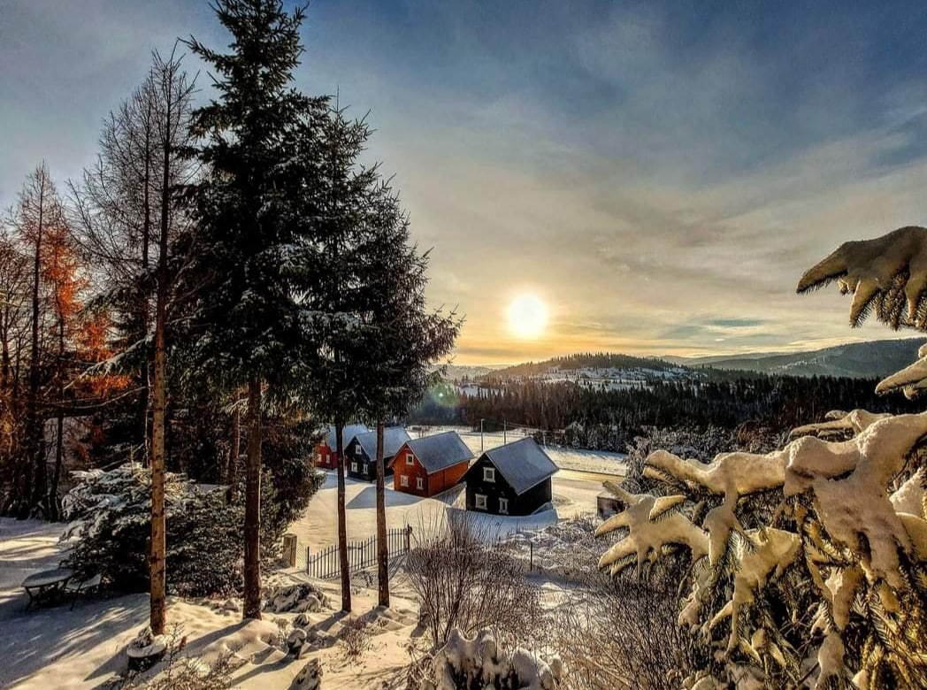 A serene winter landscape features cozy cabins nestled amid snow-covered trees. The sun rises over distant mountains, casting a warm golden light across the snowy terrain. The scene conveys a peaceful ambiance, emphasizing nature's beauty in a tranquil setting.