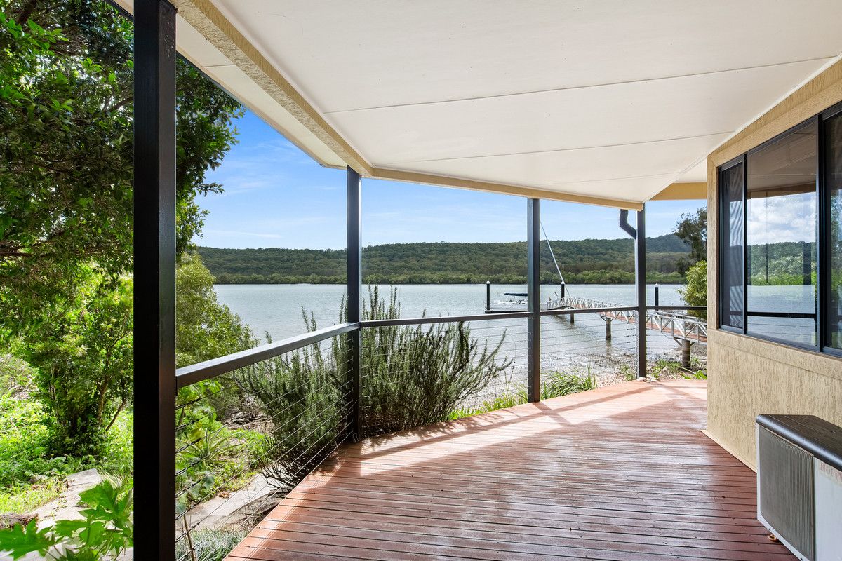 A covered deck area opens up to stunning views of the water and surrounding greenery. A wooden railing frames the space, while the natural light illuminates the deck's surface, blending indoor and outdoor living.