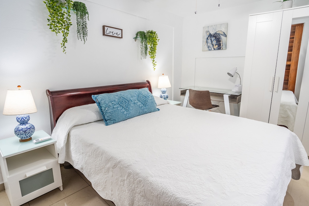 A cozy bedroom is equipped with a spacious bed covered in a white quilt. A decorative blue pillow adds a pop of color. Two bedside lamps illuminate the space, while plants hang softly on the walls. A workspace with a desk and chair is situated in the corner.