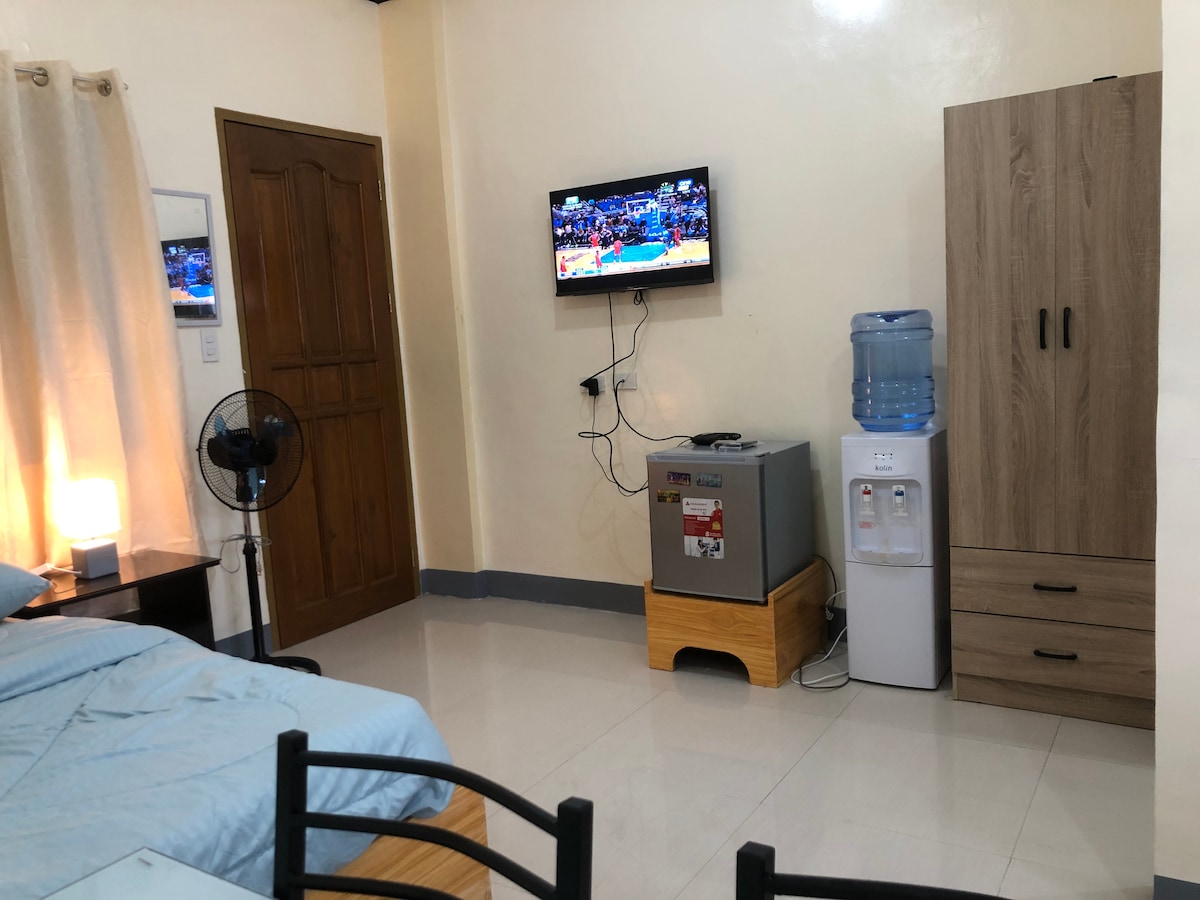A studio apartment features a comfortable bed with light blue bedding, accompanied by a small table and chairs. A wall-mounted television displays a program, while a water dispenser and a mini fridge are positioned beside a wooden cabinet. Natural light filters through light curtains.