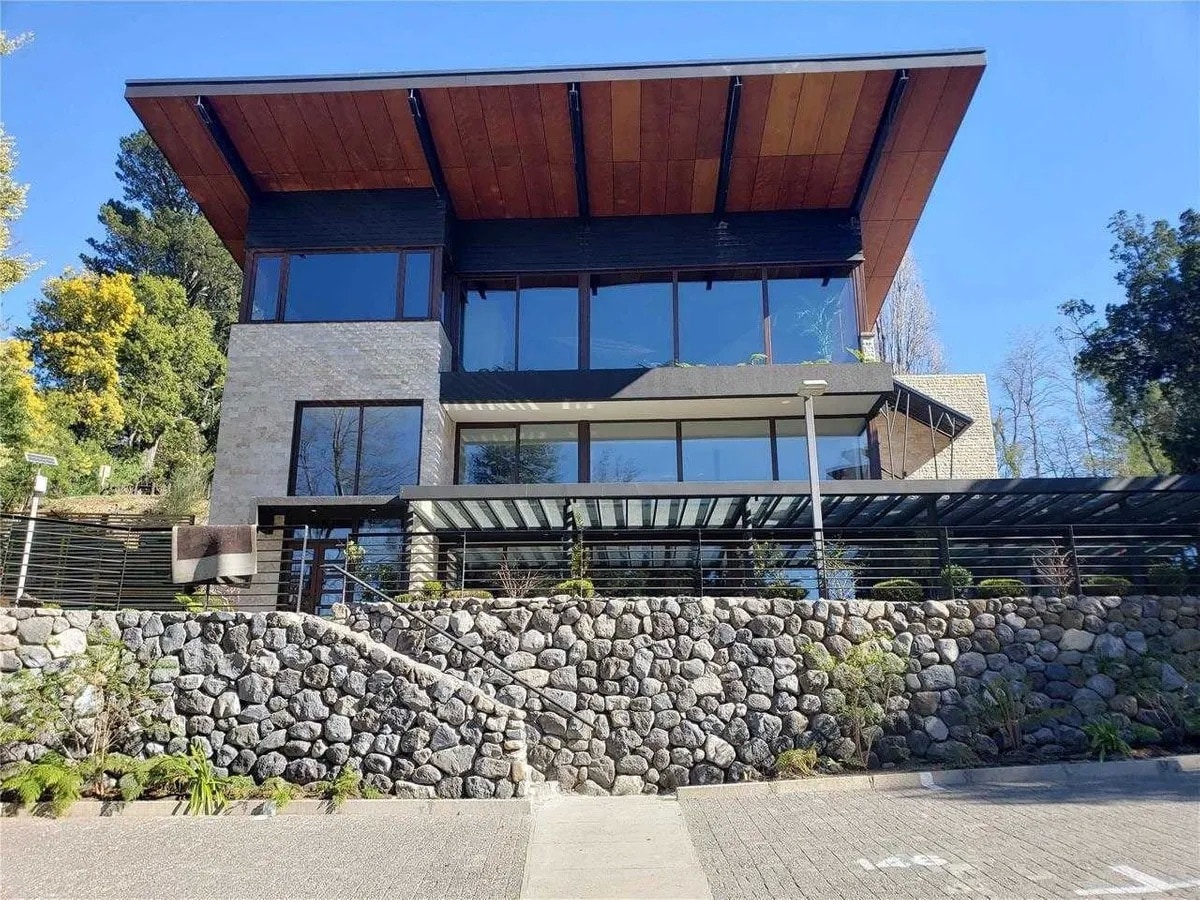 The modern building exhibits a blend of stone and glass, framed by wooden accents on the roof. A landscaped area with a stone wall and greenery enhances the entrance. Large windows allow natural light to fill the interior, offering views of the surrounding environment.