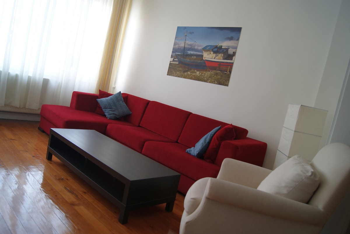 A spacious living area features a large red 'L' shaped sofa complemented by two accent pillows. A neutral-colored armchair and a dark coffee table are positioned on wooden parquet flooring. Natural light is drawn in through floor-to-ceiling curtains, illuminating a wall adorned with a coastal-themed artwork.