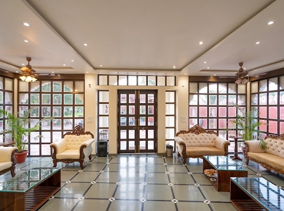 A spacious living area is furnished with elegant sofas and glass-topped coffee tables. Large windows allow natural light to fill the room, creating an airy ambiance. Potted plants add a touch of greenery, while ceiling fans provide a cooling effect.