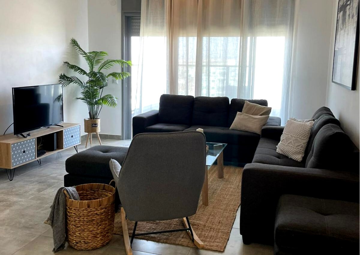 A modern living area features a spacious gray sectional sofa positioned around a glass coffee table. A plush armchair complements the seating arrangement. Light filters through sheer curtains, and a potted plant adds a touch of greenery. A woven basket rests on the floor, enhancing the decor.