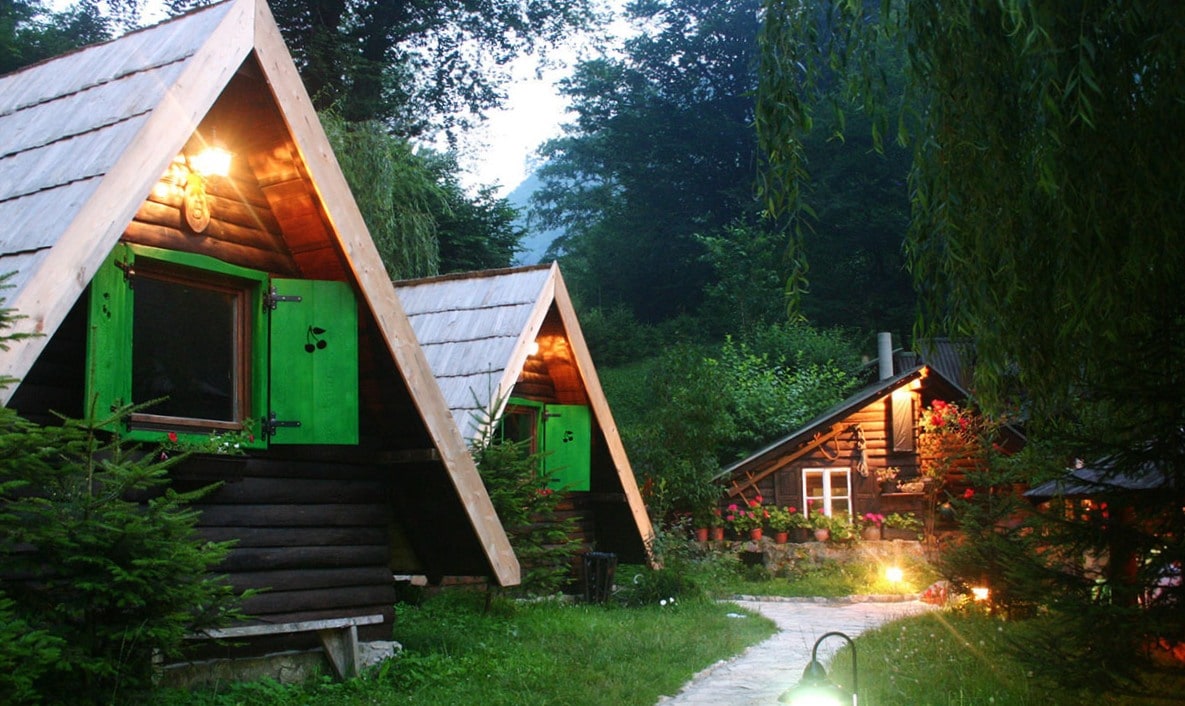 Two charming cabins with triangular roofs are situated amidst lush greenery. Vibrant green window frames contrast with the dark wooden structure. A stone pathway, illuminated by soft lighting, leads to the cabins, creating an inviting and serene outdoor space.
