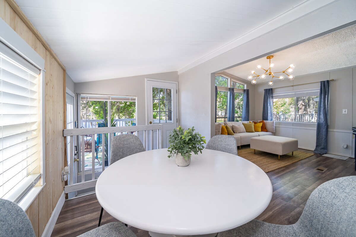 A circular dining table is surrounded by four upholstered chairs, creating a welcoming space. In the background, large windows allow natural light to illuminate the room, while a modern light fixture hangs overhead. A comfortable seating area is visible, framed by large windows and soft curtains.