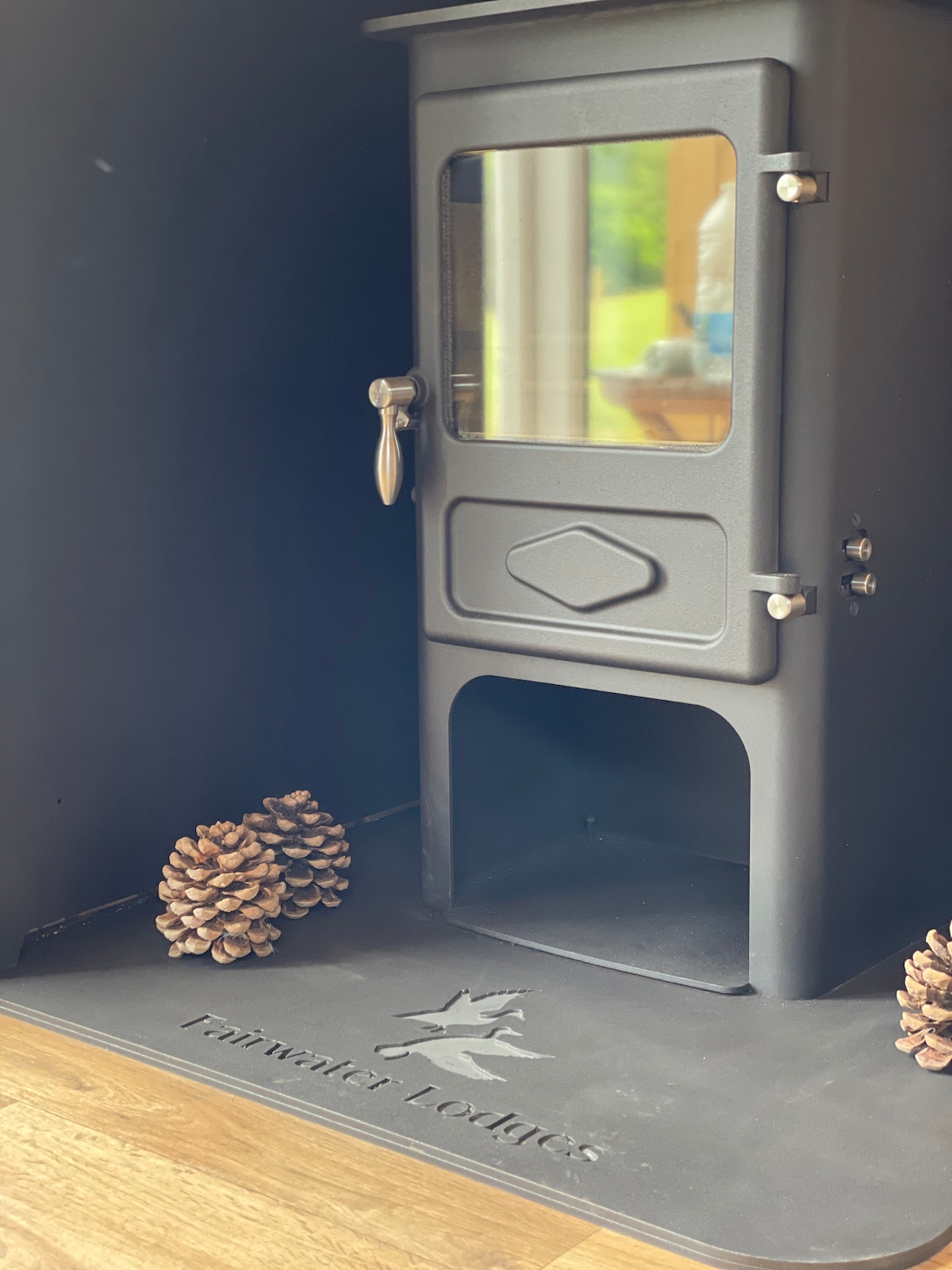 A wood-burning stove is positioned within a cozy lodge setting, featuring a glass front that allows for a view of the flames inside. Two pine cones are placed on a mat embossed with 'Fairwater Lodges,' enhancing the rustic charm of the space.