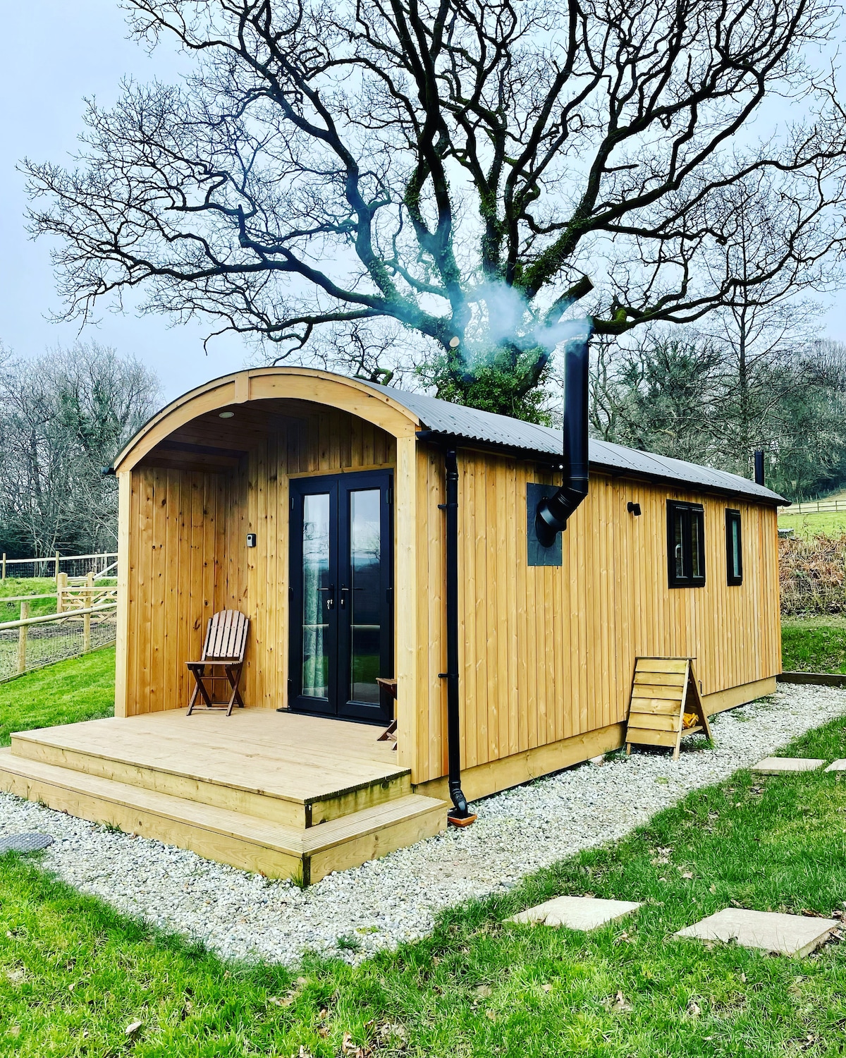 A wooden lodge with a curved roof is set amidst lush greenery. The exterior features large glass doors that open to a small deck area. A chimney emits a light plume of smoke, suggesting warmth from an interior log burner.