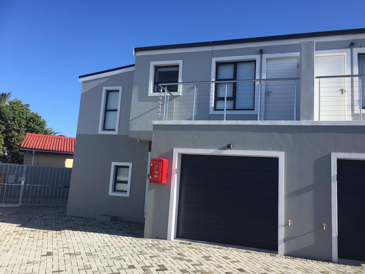 The exterior of a modern townhouse features a gray facade with large windows and a balcony, framed by a fenced yard. A red mailbox stands beside the garage door, which provides access for parking. Clear blue skies create a bright atmosphere.