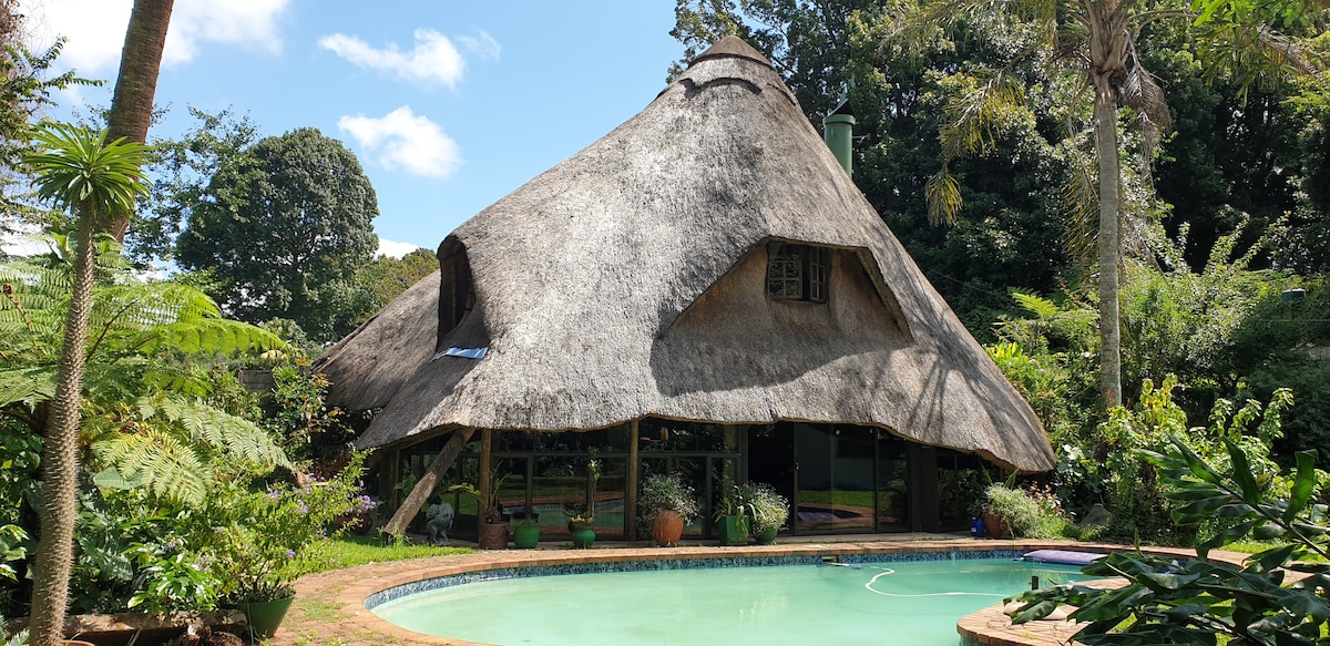The Lapa features a distinctive thatched roof structure set amidst lush greenery. A sparkling pool is positioned in front, surrounded by a well-maintained garden. The building's natural materials blend harmoniously with the surrounding tropical landscape.