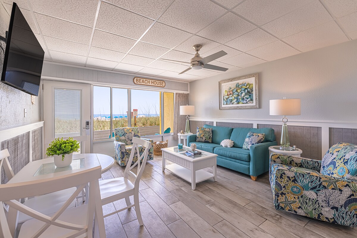 A bright living area features a turquoise couch and two patterned chairs, complemented by a white coffee table. A round dining table for four occupies one corner. Large windows allow natural light and offer views of the beach, enhancing the space's airy feel.