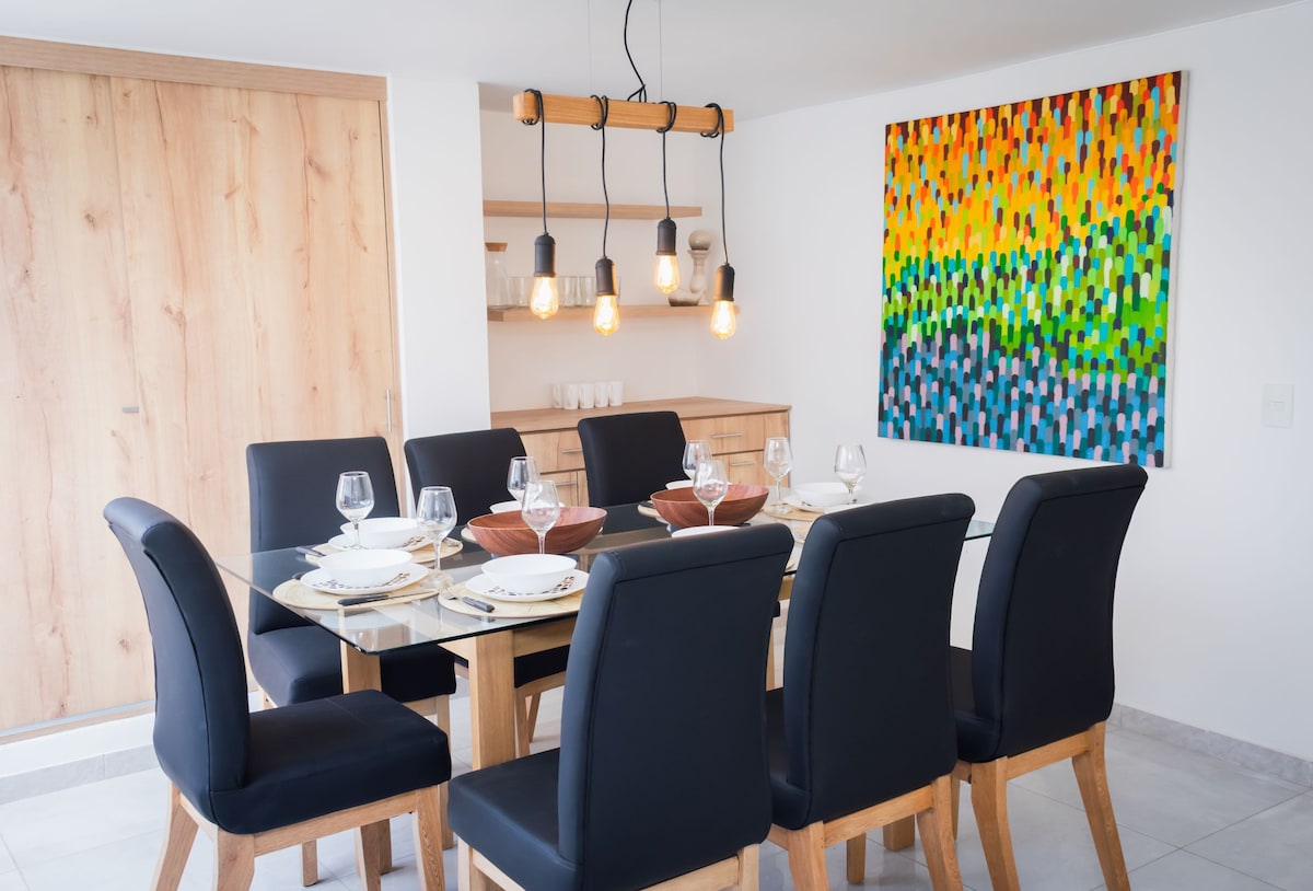 A dining area is presented with a large glass table surrounded by six black upholstered chairs. A striking piece of artwork in vibrant colors is displayed on the wall. Natural wood accents are featured in the cabinetry and light fixtures above, providing a modern yet inviting environment.