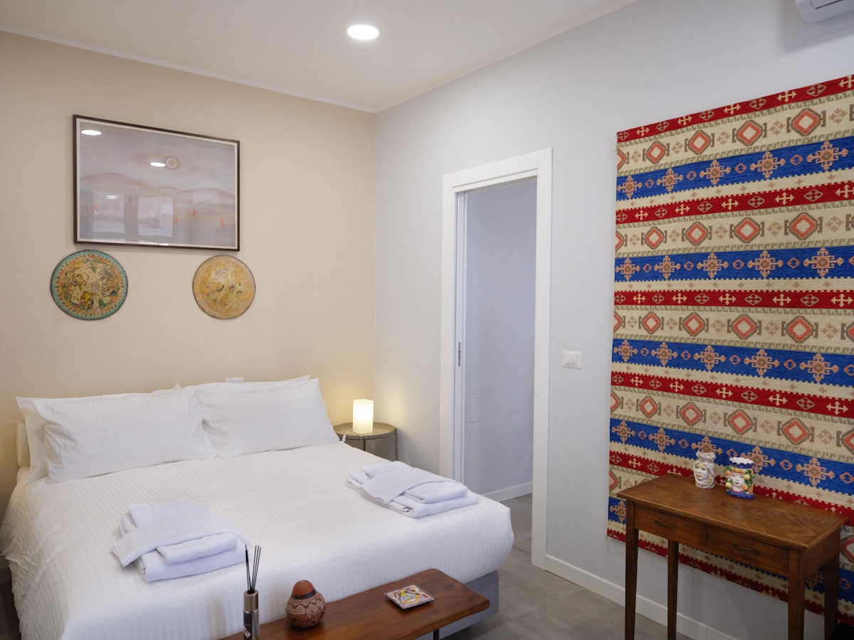 A cozy bedroom is featured, showcasing a spacious bed with crisp white linens and two neatly arranged towels on top. A small wooden table is positioned beside the bed, while decorative art and a patterned fabric panel add visual interest to the walls.