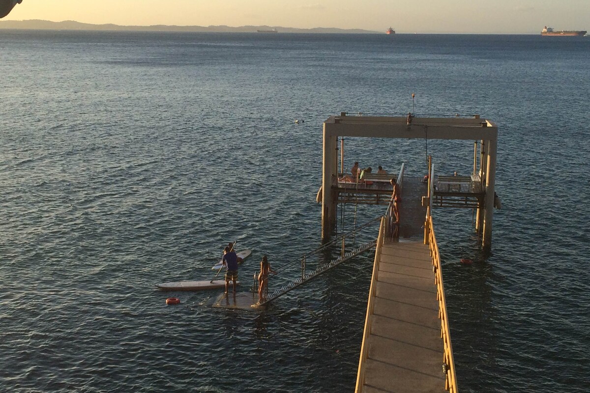 A pier extends over clear water, with several individuals engaged in activities on paddleboards. Soft waves gently lap at the structure, while distant ships can be seen on the horizon. The scene captures the tranquil ambiance of the waterfront.