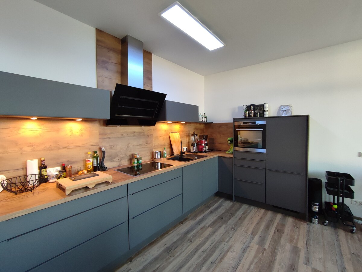 The modern kitchen features sleek grey cabinetry and elegant wooden accents. A built-in cooktop is positioned beneath a stylish range hood, with under-cabinet lighting highlighting the workspace. A refrigerator and countertop appliances are visible, alongside organized storage for spices and cooking essentials.