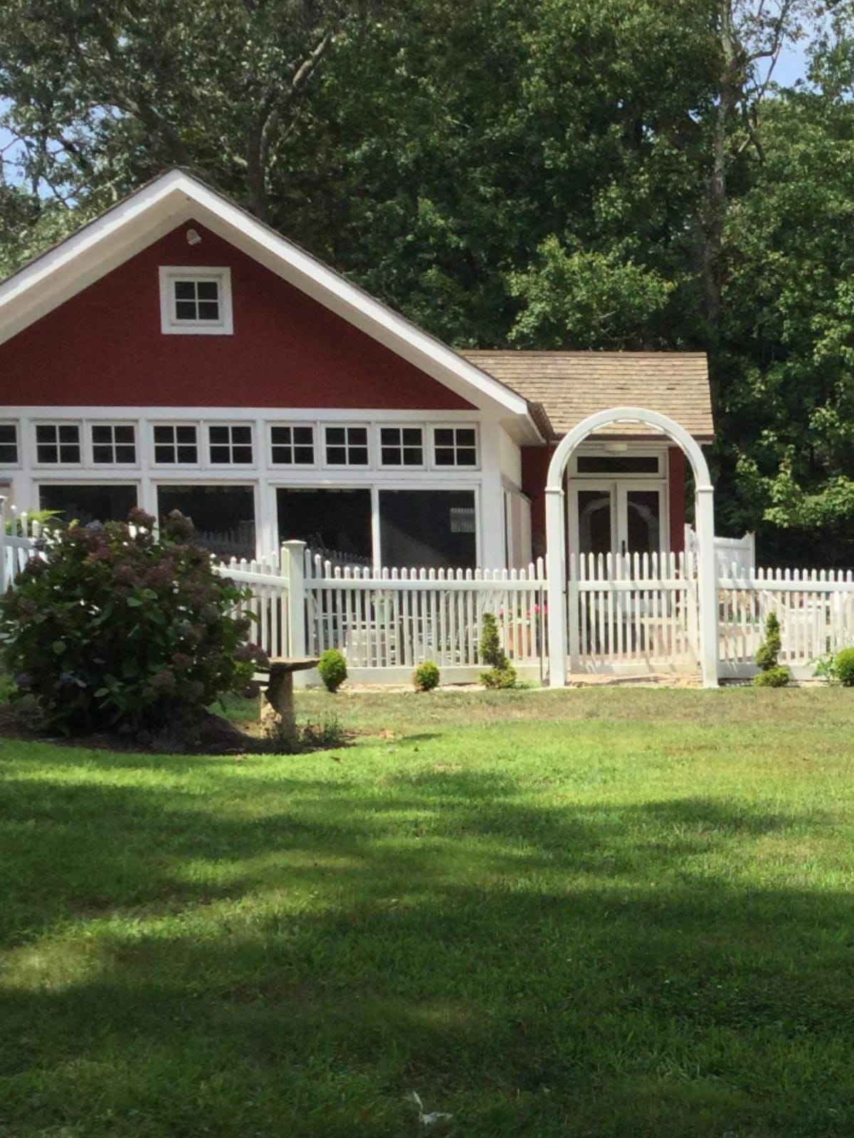 A charming red cottage is framed by a white picket fence, surrounded by a well-maintained lawn dotted with shrubs and small trees. Large windows offer a glimpse of the inviting interior, while a curved entryway draws attention to the welcoming facade.