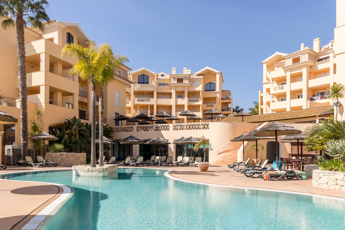 A spacious outdoor pool is surrounded by lounge chairs, tropical plants, and palm trees. Sun umbrellas provide shade, while the multi-storied resort buildings rise in the background, showcasing a harmonious blend of architecture and nature.