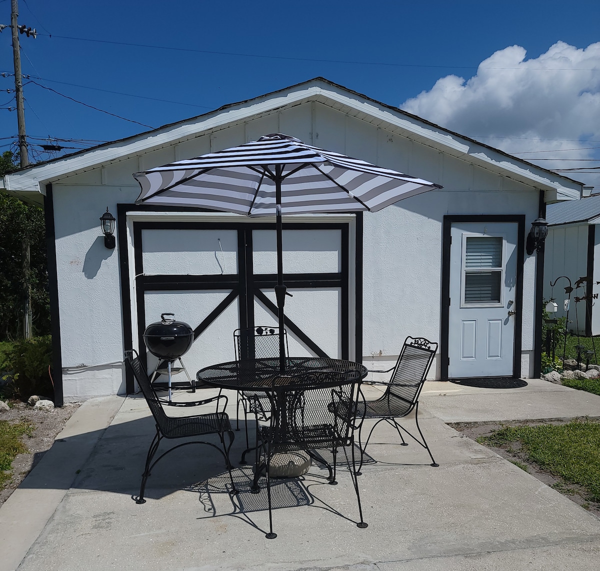 An outdoor dining area features a round table surrounded by four wrought iron chairs, shaded by a striped umbrella. A charcoal grill stands nearby on a concrete patio, framed by a simple, white building with black accents and a door.